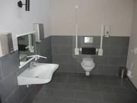 Toilettenraum mit Behindertentoilette rechts und links Handläufe und links ein Waschbecken mit Spiegel