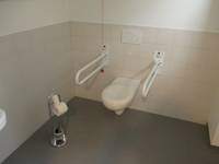 An einer hellen Wand eine weiße Toilette mit Haltegriffe auf beiden Seiten