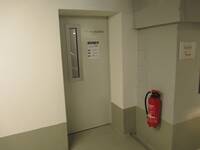 Eine helle Türe mit tiefer Türleibung in einer weißen Wand. Rechts von der Tür hängt ein Feuerlöscher.