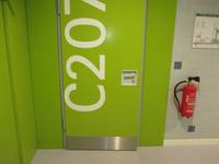 eine grüne Tür mit großen weißer Aufschrift C207, rechts daneben hängt ein Feuerlöscher an der Wand