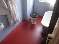 Badezimmer mit einem roten Boden. Rechts ist eine  freistehende Badewanne. Am Ende des Raums steht schräg zur Wand eine Toilette