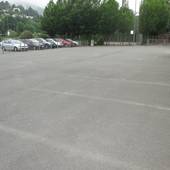 Ein großer asphaltierter Platz, links ein angrenzender Parkplatz, durch Metallgitter abgetrennt. Im Hintergrund Bolzplatz und hohe Bäume