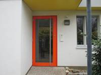 überdachte Glaseingangstür mit orangefarbenen Metallrahmen und senkrechter Metallstange, rechts davon Klingel und Fenster