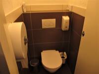 Eine weiße Toilette an einer dunklen Wand. Links neben dem WC hängt ein großer runder WC-Papierspender