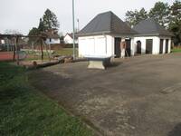 Ein großer asphaltierter Platz, von  Grünflächen umgeben. Vor zwei kleinen Lagergebäuden steht eine Tischtennisplatte. Links in der Grünfläche ist eine Nestschaukel, im Hintergrund ist ein Sportplatz.
