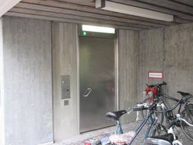 überdachter Bereich mit einer Betonwand, in der Wand einflügelige Metalltür, links davon Bedienfeld, im Vordergrund Fahrräder