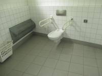 weißes Hänge-WC an einer weißen Wand. Rechts und links jeweils ein Haltegriff. Hinter der Toilette Spühltaster. Graue Bodenfliesen, an der linken Wand ist ein Heizkörper.