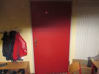 Rote Tür in einer weißen Wand, links einige Kleiderhaken mit aufgehängten Jacken und niedrigen Bänken davor