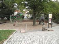 Spielplatz mit Zugang, Stufen, einem kreisförmigen Sandkasten in dessen Mitte Spielgeräte und ein Baum stehen