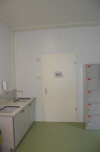 weiße Rür mit Aufschrift "WC" in hellgrauer Wand. Rechts daneben zwei Waschbecken, links daneben abschließbare Schließfächer