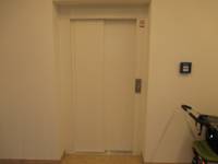 weiße geschlossene Aufzugstür,  in der Wand zurückgesetzt. Direkt neben der Tür ist eine Taste. Im Bildvordergrund rechts hängt ein Feuermelder an der Wand
