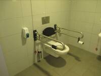 Weiße Behindertentoilette an einer deckenhoch gekachelten Wand, rechts und links vom WC Haltegriffe vorhanden