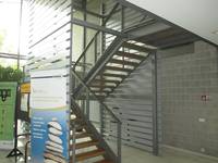 Eine freistehende Treppe aus Metall mit offenen Holzstufen, seitlich über die gesamte Höhe Glaswände mit aufgeklebten Streifen. Auf der linken Glaswandsteht ein Roll-Up und dabneben eine Pinwand