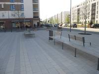 ein Platz aus Betonverbundsteinen, mit zwei längeren Bänken die parallel zu zur rechts verlaufenden Straße aufgestellt sind, im Hintergrund mehrere hohe Gebäude