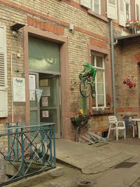 offene Eingangstür mit Rampe davor, links davon ein Geländer, rechts ein Fahrradständer und im Hofeck ein Tisch mit Stühlen