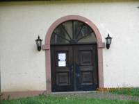 Zweiflügelige hölzerne Eingangstür, über der Tür Verglaster Bogen, rechts und links eine Lampe an einer hellen Wand