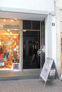 im Vordergrund Hauptstraße mit Kleinsteinpflaster und darauf folgend eine Stufe mit Podest, eine Frontalaufnahme der Glastür des \'Eingangs zur Galerie Julia Phillipi, links ein erhelltes Schaufenster 