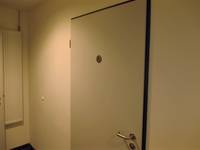 weiße Tür mit dunklem Rahmen in einer weißen Wand, auf der Tür ein Rollstuhlsymbol, links an der Wand hängt ein Heizkörper 
