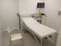 Raum mit einer Untersuchungsliege, einem Stuhl davor, rechts hinten EKG-Gerät mit Bildschirm. An der Rückwand ist eine Tür 