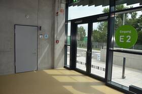 In der linken Bildhälfte Betonwand mit eingebaute grauer Tür in dunklem Metallrahmen. An der Wand rechts von der Tür hängt ein Schild mit Rollstuhlsymbol, daneben auf gleiche Höhe ein Schild mit dem Logo von "Toilette für alle. Senkrecht zur Betonwand rechts ist eine hohe, breite Glasfront mit zweiflüglicher Glaseingangstür. Rechts daneben klebt auf der Scheibe ein großer grüner Kreis mit weißer Beschriftung: Eingang E 2"