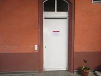 weiße Tür in einer roten Wand. Darüber Rundbogen-Oberlicht. Rechts neben der Tür steht ein Blumentopf auf dem Boden