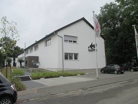 zweistöckiges Gebäude mit Schrägdach, an der Seitenwand ist der Schriftzug "Amici", vor dem Gebäude Straße, Partkplätze und eine Fahne