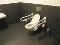 Eine weiße Toilette in einer schwarz gekachelten Wand. Auf beiden Seiten ein Haltegriff.