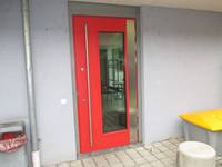 Rote Tür mit einr eingefasster Glasscheibe in hellgrau getrichener Wand. Links Regenablaufrohr und Mülleimer. Rechts Steinpoller und Streugutbehälter
