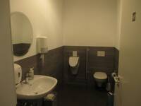 WC-Raum mit dunklem Boden und einer halbhohen dunkel gekachelten Wand.
Zusätzlich zur Toilette befindet sich ein Pissoir und ein Waschbecken im Raum.