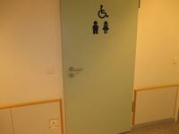 Helle Tür in einer weißen Wand. Auf der Tür sind Symbole für Mann, Frau, Rollstuhl