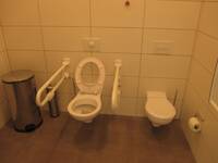Eine weiße Toilette an einer weiß gefliesten Wand. Auf beiden Seiten ist ein Haltegriff. rechts neben der Behindertentoilette ist eine Kindertoilette.
