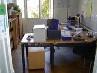 Schreibtisch mit Büroartikeln und Computerbildschirm, hinter dem Tisch steht ein Bürostuhl, der Bodenbelag ist Holzparkett  
