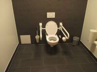 WC_Raum, der Boden ist dunkelgrau gefliest, und hintere Wand mit der weißen Toilette ist dunkelgrau gestrichen, die übrigen Wände sind weiß. Neben der Toilette sind links und rechts heruntergeklappte  Haltegriffe, am rechten Haltegriff hängt eine Rolle WC-Papier.