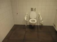 Eine weiße Toilette an einer weiß gekachelten Wand. Der Boden ist dunkel. An beiden Seiten der Toilette ist ein Haltegriff.
