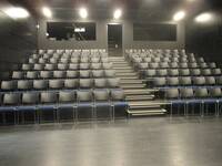 Theatersaal mit ansteigenden Sitzreihen, die auf der linken und auf der rechten Seite der Treppe angeordnet sind. Pro Seite gibt es 7 Sitzreihen mit je 8 Stühlen. Die Stühle haben eine graue Rückenlehne und eine blaue Sitzfläche. Die Füße der Stühle bestehen aus Metall. 