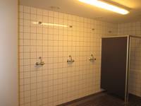  weiß gekachelte Wand mit drei Duschplätzen, links an der Wand eine abgetrennte Dusche, dunkle Bodenfliesen
