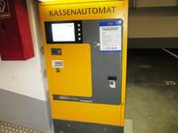 Kassenautomat links oben Bildschirm mit einer Reihe senkrechten Bedienknöpfe daneben, darunter 2 Karteneingabeschlitze noch weiter unten Ausgabefach, rechts Münzeinwurf und Geldscheineingabe  