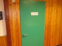 Grüne Tür in einer mit Holzpaneelen verkleideten Wand. Auf dem Türschild steht WC, daneben Symbol für Rollstuhlfahrer, Mann und Frau
