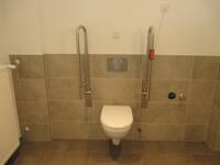 Eine weiße Hängetoilette in einer halbhoch hellbraun gefliesten Wand. Rechts und links von der Toilette sind hochgeklappte Haltegriffe