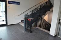 links Tür zu Foyer, daneben Treppe, die aufwärts und nach unten führt. Rechts von der Tür zum Foyer hängt ein blaues Schild mit der weißen Beschriftung "Lounge, Logen" und einem Treppensymbol. Rechst neben der abwärts führenden Treppe ist der Aufzug