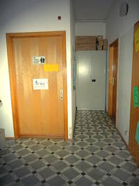 Links die geschlossene Tür zur Behindertentoilette, auf der Tür ist ein Hinweisschild für die Behindertentoilette, ansonsten ist da der Flur, 2 Türen zu den Büroräumen und ein Schrank