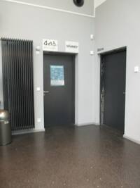 Raumecke mit einer schwarzen Tür links zu Männer - und Behindertentoilette. Links neben der Tür hängt ein langer, schwarzer Heizkörper. Rechts in der anderen Raumecke ist eine weitere schwarze Tür.