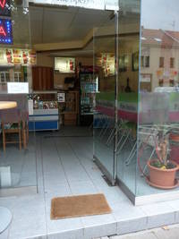 Offene Glastür mit einer Stufe davor , im Eingang Schmutzabweiser, im Hintergrund der Laden mit Theke und rechts Stühle 