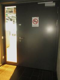 Die Tür und der Türrahmen sind schwarz. Direkt links von der Tür ist eine bodentiefe Glasfläche. Auf der Tür ist Schild im DIN A4-Format mit dem Symbol für "Rauchen verboten" angebracht
