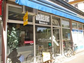 gläserne Ladenfront mit Tür und Schaufenster darauf Schriftzug "Schlüsseldienst Marschollek", darüber eingezogene blaue Markisse, davor zwei abgestellte Fahrräder