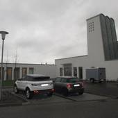 L-förmiges Flachdachgebäude mit einem Turm auf der rechten Seite, davor ein Platz und an der Straße Parkplätze mit Autos