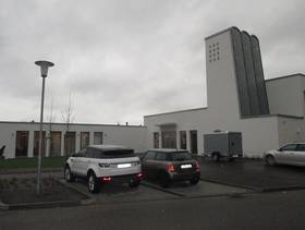L-förmiges Flachdachgebäude mit einem Turm auf der rechten Seite, davor ein Platz und an der Straße Parkplätze mit Autos