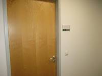braune Holztür in einer weißen Wand, rechts neben der Tür Schild mit Raumbezeichnung