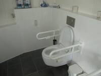 Ein weißes Hänge WC an einer weiß gekachelten Wand. Der Boden ist dunkel gefliest. Auf beiden Seiten sind weiße Haltegriffe