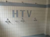 ein gekachelter Raum mit dem Schriftzug \"HTV\" in blauen Kacheln, im Raum drei Duschplätze